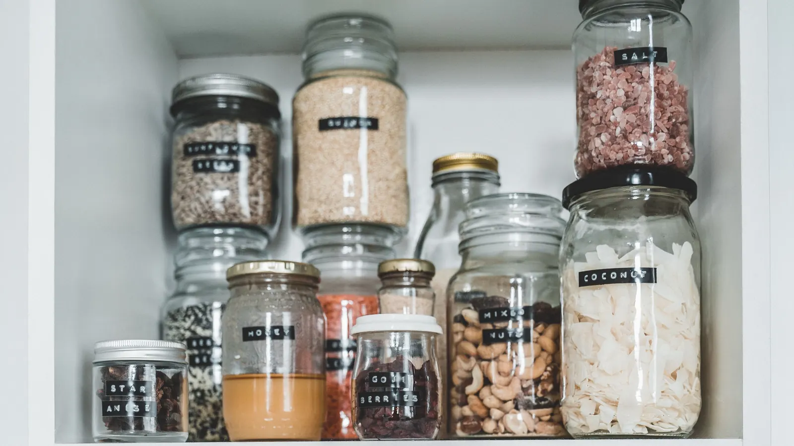Labelled glass jars of nuts, spices and other ingredients on a white cupboard shelf