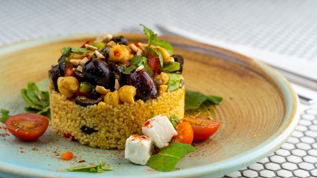 A plated couscous salad tower topped with chickpeas, olives, fresh herbs and chopped nuts, served with feta cubes and cherry tomatoes.