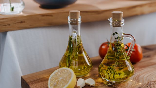 Two glass cruets of herb-infused oil on a wooden board with a halved lemon, garlic cloves, and fresh tomatoes.