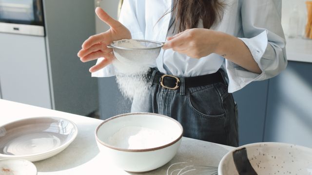 A person sieving white flour into a ceramic bowl on a kitchen countertop.