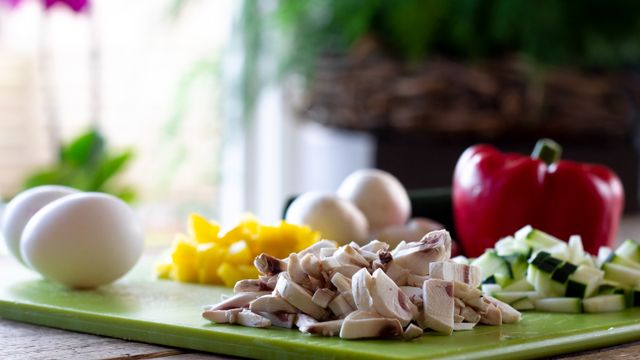 Chopped button mushrooms, diced courgette and yellow pepper arranged on a green cutting board next to a whole red pepper and eggs