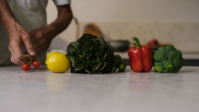 A person placing fresh cherry tomatoes alongside a lemon, red pepper, broccoli, and salad greens on a kitchen worktop