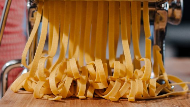 Freshly cut ribbons of pasta dough emerging from a metal pasta machine onto a wooden surface.