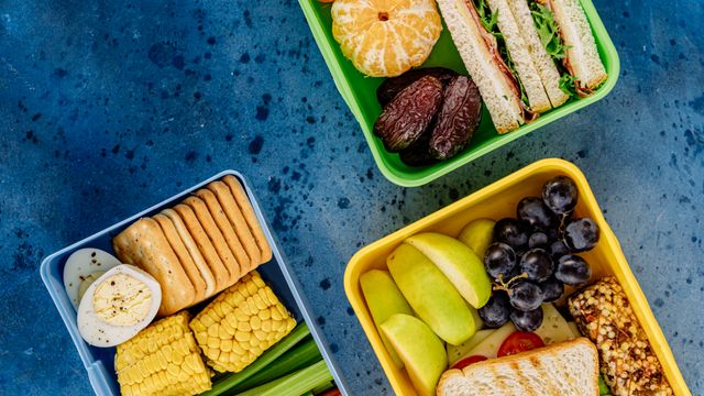 Top-down view of three colourful lunchboxes filled with sandwiches, fruit, boiled egg, crackers and vegetables.