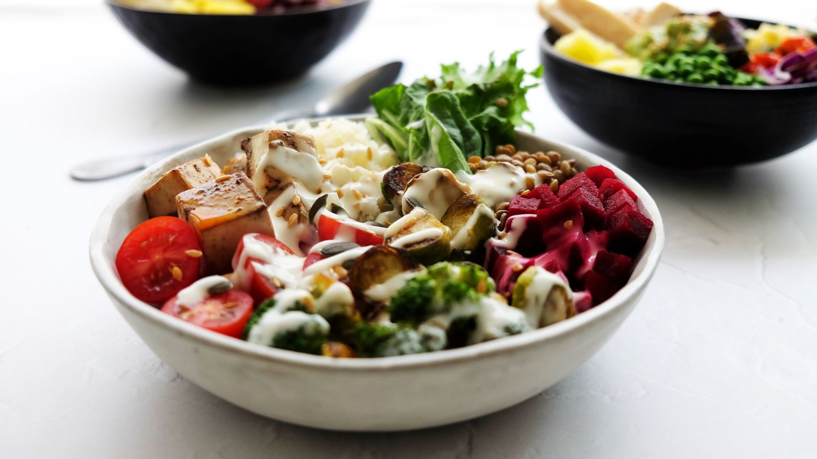 A lunch bowl filled with grains, broccoli, cherry tomatoes, beetroot, tofu cubes, and a drizzle of dressing.