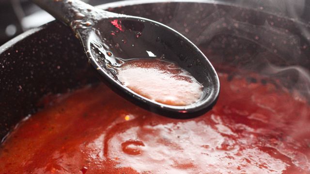 Simmering red tomato sauce in a cooking pan with a black spoon held above it