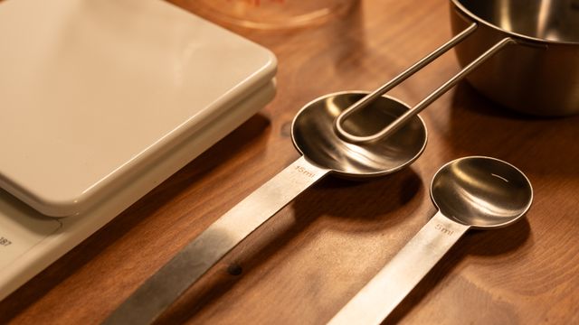 Metal measuring spoons and a measuring cup next to a white kitchen scale on a wooden table.