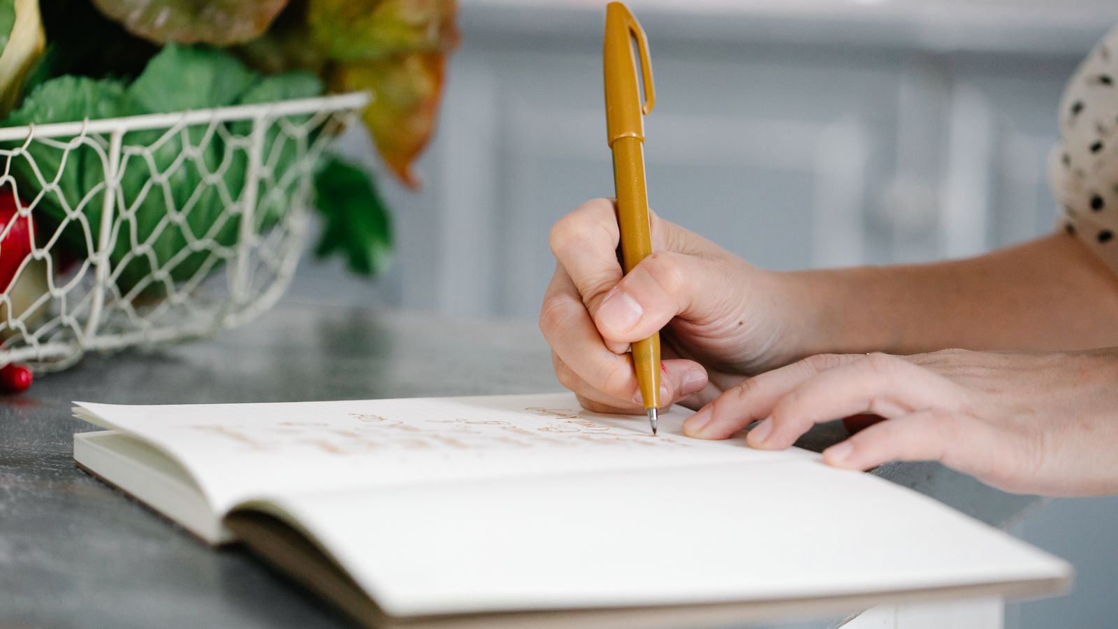 A person writing with a pen in an open notebook on a kitchen counter near a basket of fresh produce.