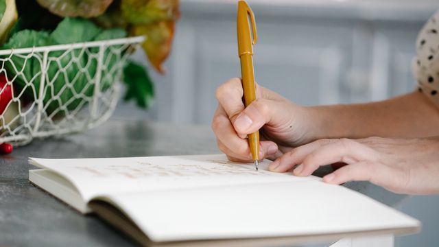 A person writing with a pen in an open notebook on a kitchen counter near a basket of fresh produce.