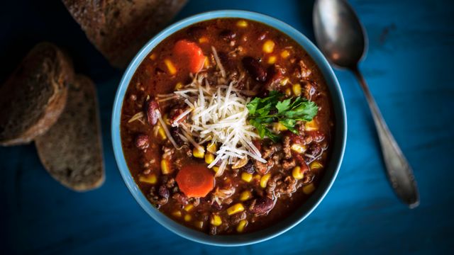A bowl of hearty bean and beef stew garnished with grated cheese and fresh herbs, served on a blue table alongside crusty bread.