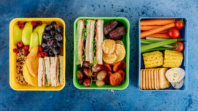 Three colourful lunchboxes packed with sandwiches, fresh fruit, crackers, and vegetable sticks on a blue surface.